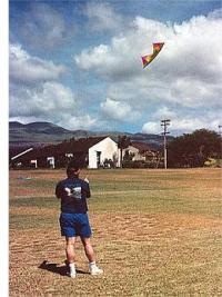 This man is flying a colorful quad kite in a grassy vacant block.